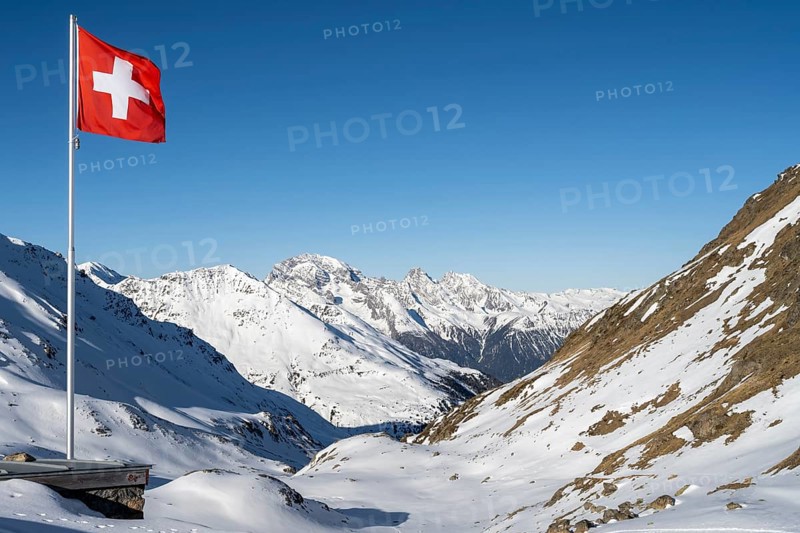 Swiss flag in front of mountain landscape in winter, mountain panorama with the peaks of the Bergüner Stöcke Tinzenhorn and Piz Ela, view at the Keschhütte in winter, Bündner Haute Route, Albula Alps, Rhaetian Alps, Grisons, Eastern Switzerland, Switzerland, Europe