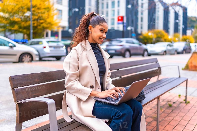 African american elegant businesswoman sitting on a bench using laptop in the city in autumn