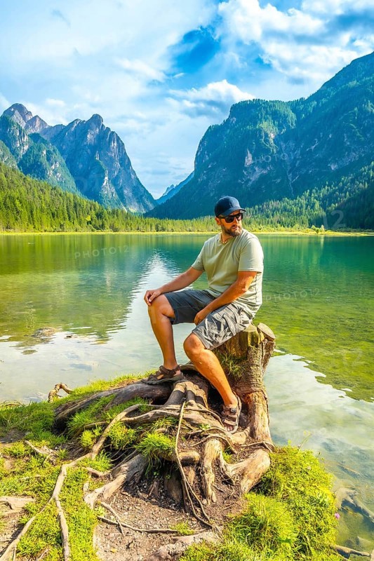 Tourist enjoying the scenic view of lake dobbiaco and the dolomites mountains while sitting on a tree stump by the water