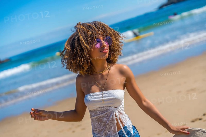 Happy brunette beauty woman wearing summer clothes, sunglasses and holding a sun hat on hand standing on the beach enjoying summer