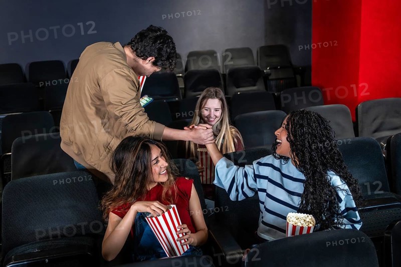 Friends enjoying popcorn and shaking hands while watching an exciting movie in a lively cinema, sharing smiles and laughter together