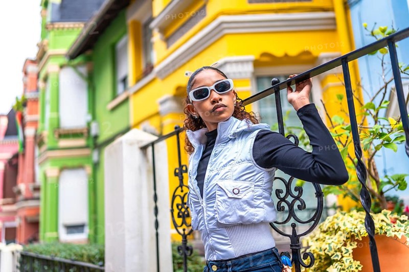 African american female model leaning on a rail with trendy futuristic clothes and sunglasses next to colorful houses
