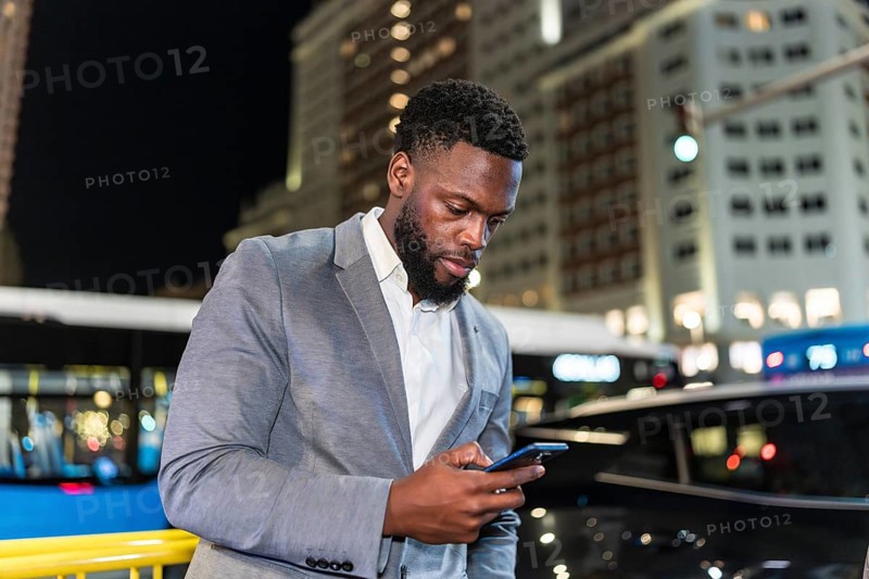 Young businessman using smartphone on a city street at night, with office buildings and traffic in the background