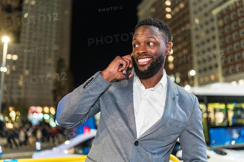 Young businessman smiling and chatting on a smartphone in a vibrant urban city at night, surrounded by dazzling lights and modern buildings