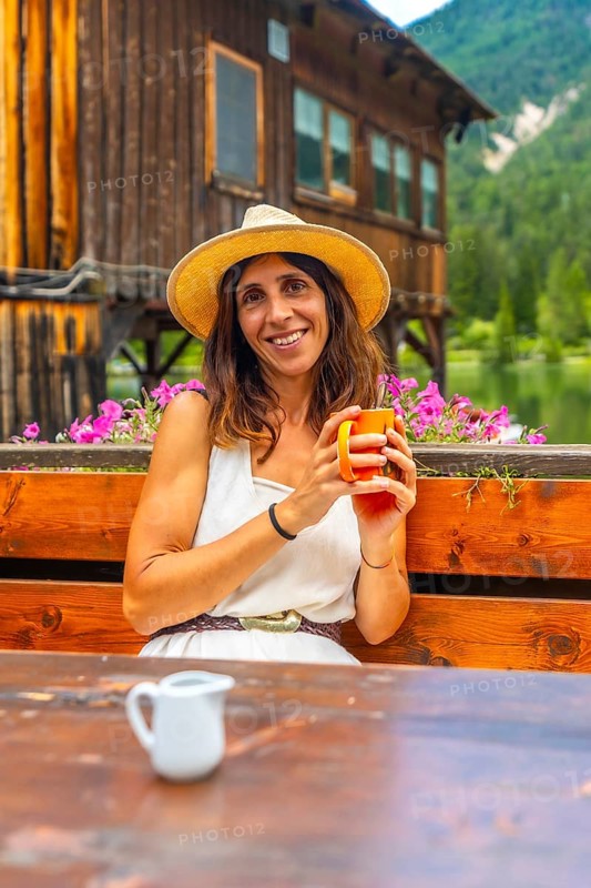 Woman wearing a straw hat enjoying a hot beverage in a mountain chalet by the lake in the italian dolomites
