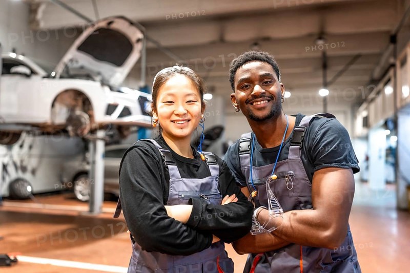 Two smiling mechanics with arms crossed, showcasing confidence and professionalism in a busy car service station filled with tools and equipment
