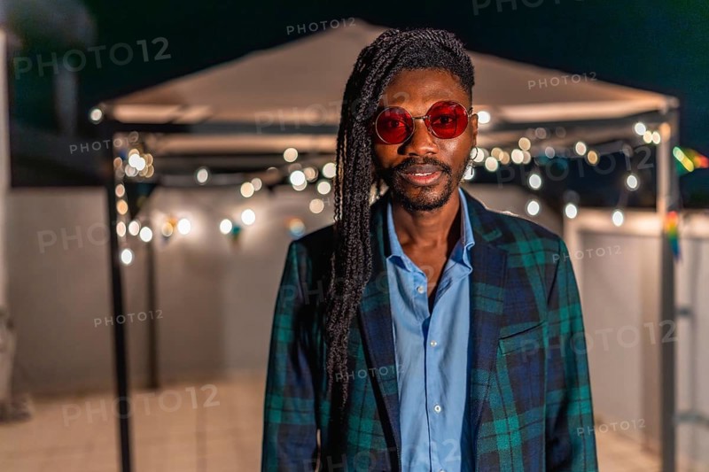Portrait of a cool african man with afro hairslyle and colorful vintage jacket and sunglasses standing at night party in a terrace