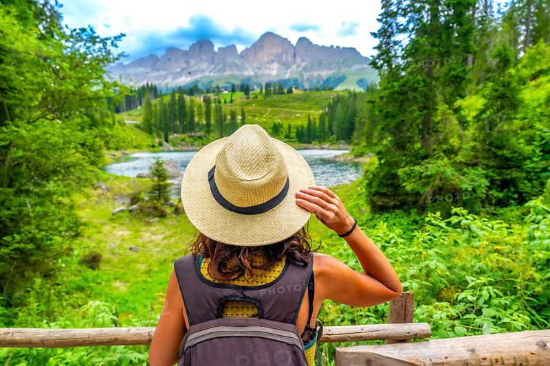 Female tourist with backpack and straw hat admiring the beautiful landscape of lake carezza and latemar mountain in the italian dolomites