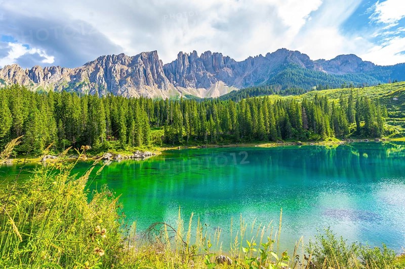 Emerald waters reflecting the latemar group and surrounding pine forest under a blue sky with clouds at karersee in summer