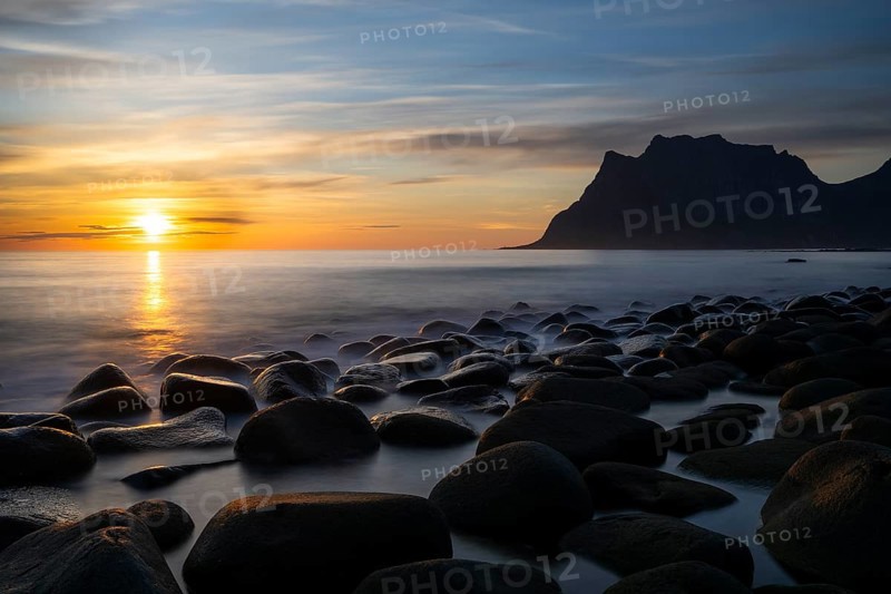 Seascape on the beach at Uttakleiv (Utakleiv), in the foreground round stones in the sea. In the background the mountain Hogskolmen. At night at the time of the midnight sun. The sun is low in the sky. Early summer. Long exposure. Uttakleiv, Vestvagoya, Lofoten, Norway, Europe