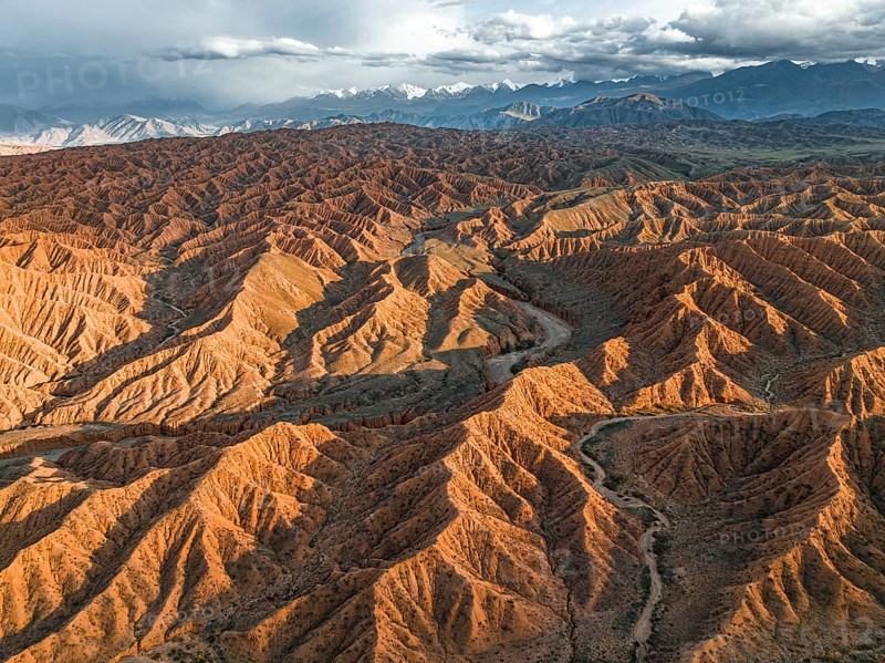 River bed runs through a landscape of eroded hills, badlands at sunset, mountain peaks of the Tian Shan Mountains in the background, aerial view, Canyon of the Forgotten Rivers, Issyk Kul, Kyrgyzstan, Asia