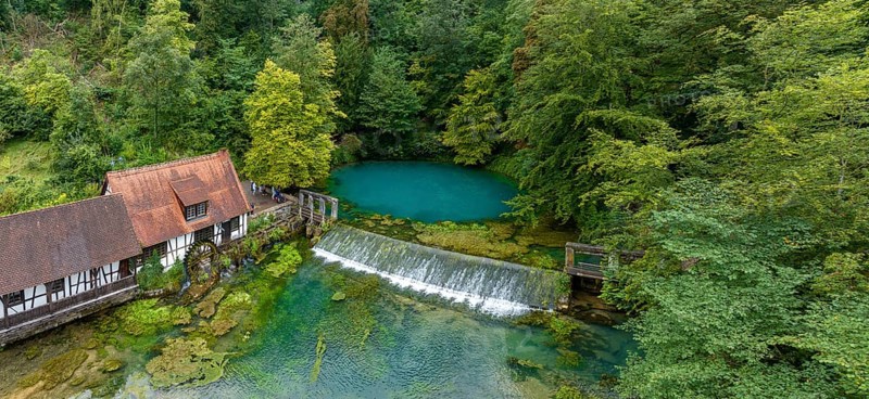 Blautopf Blaubeuren with industrial monument Hammerschmiede, source of the little river Blau in a landscape with forest. Karst spring, geotope and geopoint of the UNESCO Swabian Alb Geopark, tourist attraction. The popular excursion destination is now being thoroughly renovated and will therefore be closed to visitors until the end of 2028. Drone photo. Blaubeuren, Baden-Württemberg, Germany, Europe