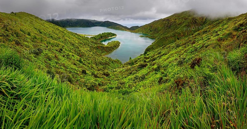 Natural landscape with a green slope and a lake surrounded by hills and a cloudy sky, crater lake Lagoa do Fogo, Caldeiras, Sao Miguel Island, Azores, Portugal, Europe