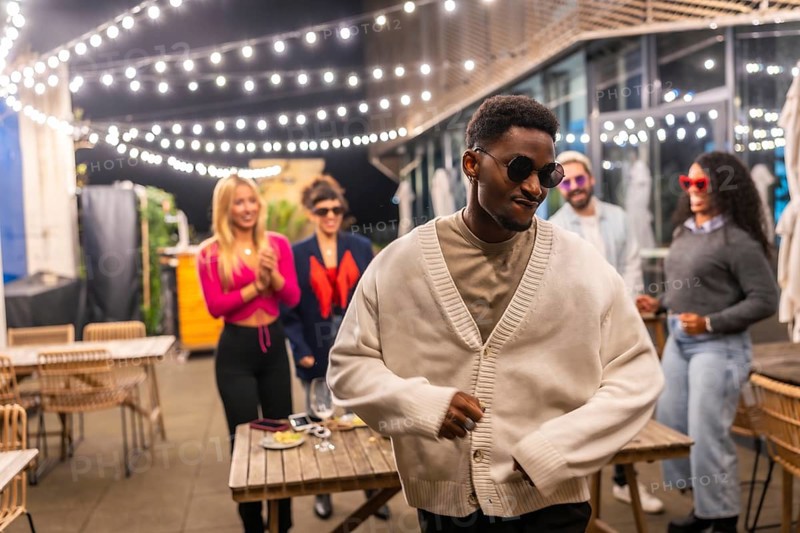 Cool african man with sunglasses and warm clothes dancing in a night roof bar party next to multiracial friends