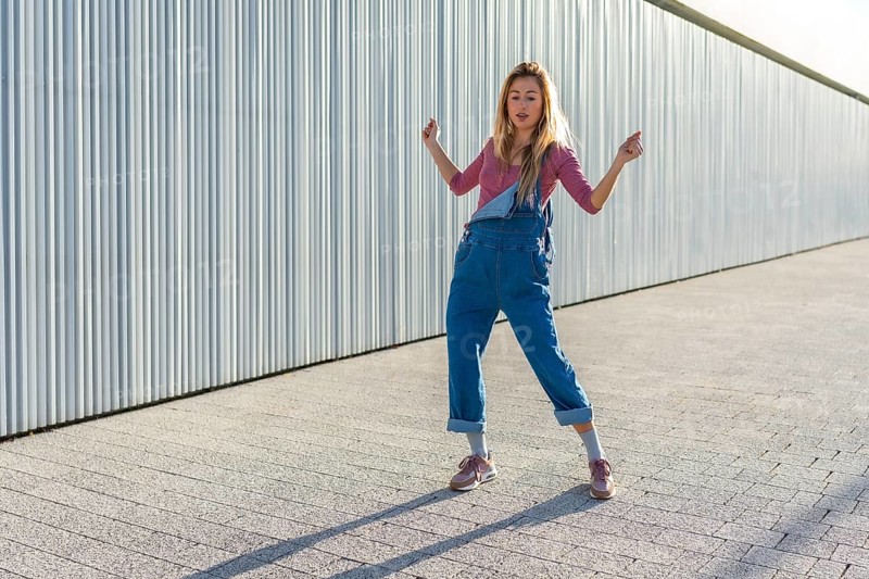 Blonde dancer performing urban dance moves on a paved area against a corrugated metal wall, showcasing energy and style