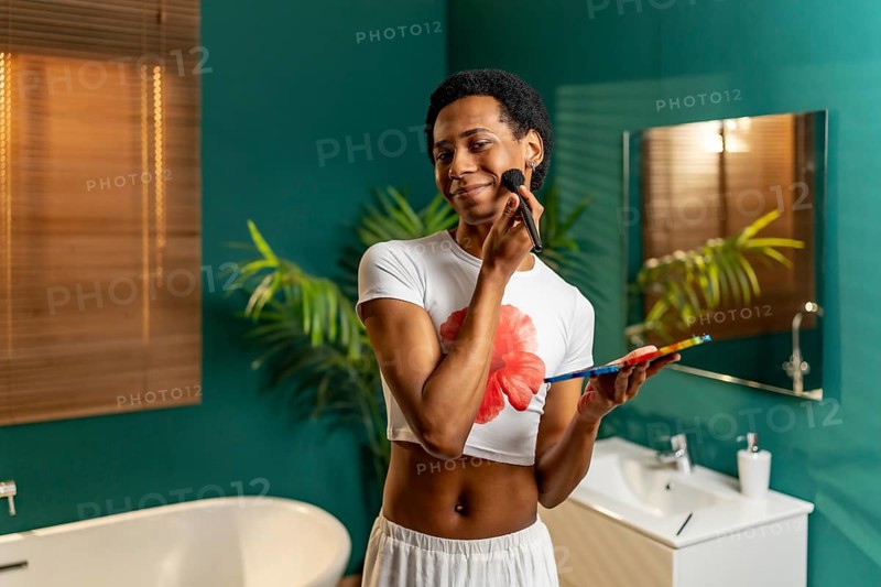 Cute smiling gay man making up at home standing in the bathroom using brush and palette