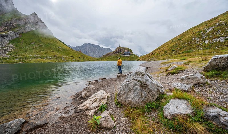 Mountaineer in yellow jacket at the mountain lake, view over the Wolayersee lake to the alpine club hut Wolayerseehütte, cloudy mountain landscape, Carnic Alps, Carnic High Trail, Carinthia, Austria, Europe