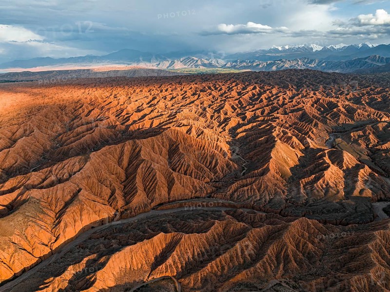 River bed runs through a landscape of eroded hills, badlands at sunset, mountain peaks of the Tian Shan Mountains in the background, aerial view, Canyon of the Forgotten Rivers, Issyk Kul, Kyrgyzstan, Asia