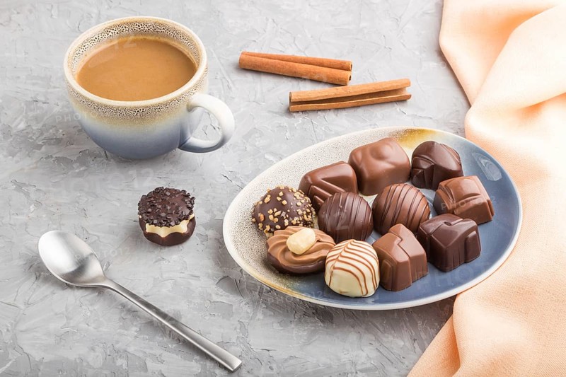 Different chocolate candies and a cup of coffee on a gray concrete background and orange textile. side view, close up
