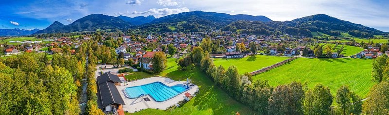 Panoramic view of a village with a swimming pool, surrounded by green meadows and forests, against a mountain backdrop, Bad Feilnbach