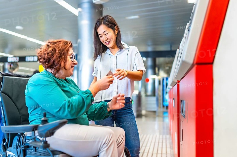Chinese smiling caregiver buying metro ticket to a caucasian adult disabled woman in wheelchair