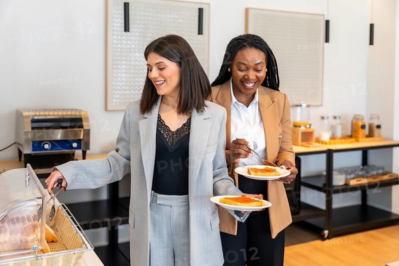 Two businesswomen are smiling while having breakfast at a hotel buffet, showing a positive and productive start to their business trip