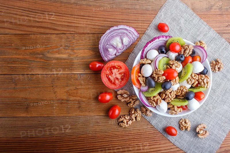 Salad with fresh cherry tomatoes, mozzarella cheese, olives, kiwi, onion and walnuts on brown wooden background, copy space, top view