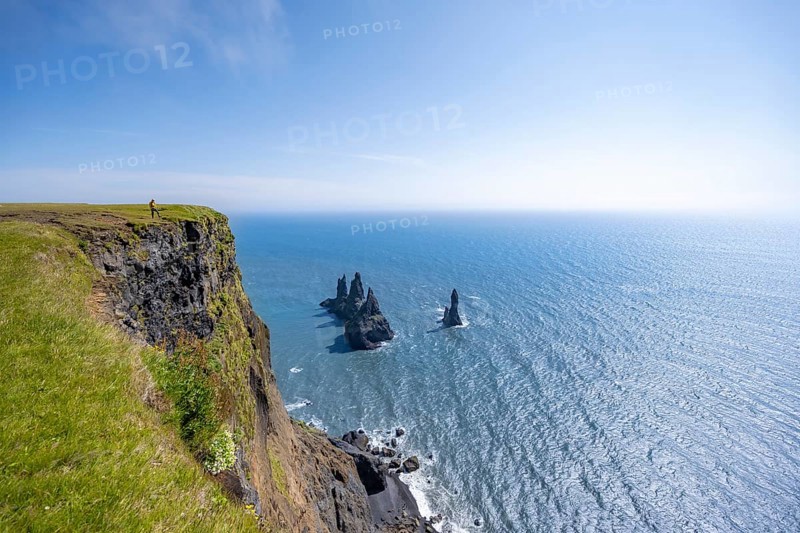 Young man standing on cliff, view from steep cliff to the rocks Reynisdrangar in the water, at Reynisfjara beach, Vik, South Iceland, Iceland, Europe