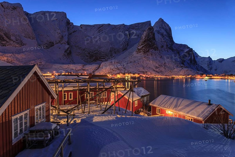 Red wooden houses on a fjord in front of snow-covered mountains, illuminated, twilight, winter, Reine, Moskenesoya, Lofoten, Norway, Europe