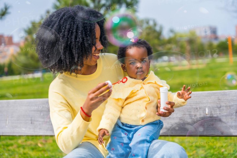 African girl on lap of her mother playing soap bubbles sitting on a park bench
