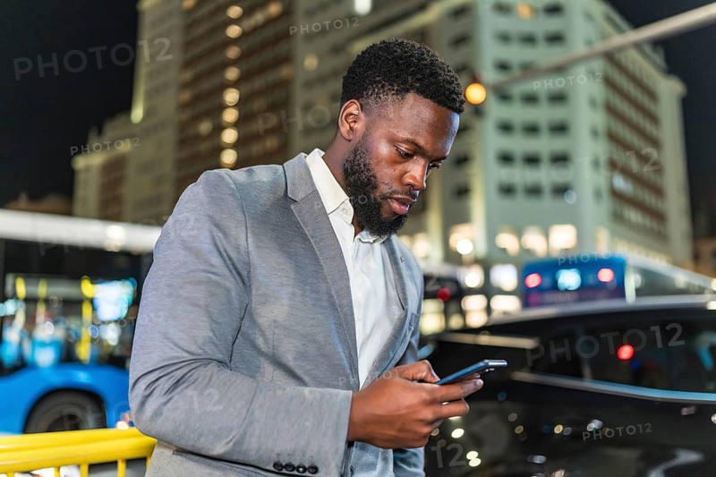 Young businessman using smartphone on city street at night, with blurred buildings and traffic in background