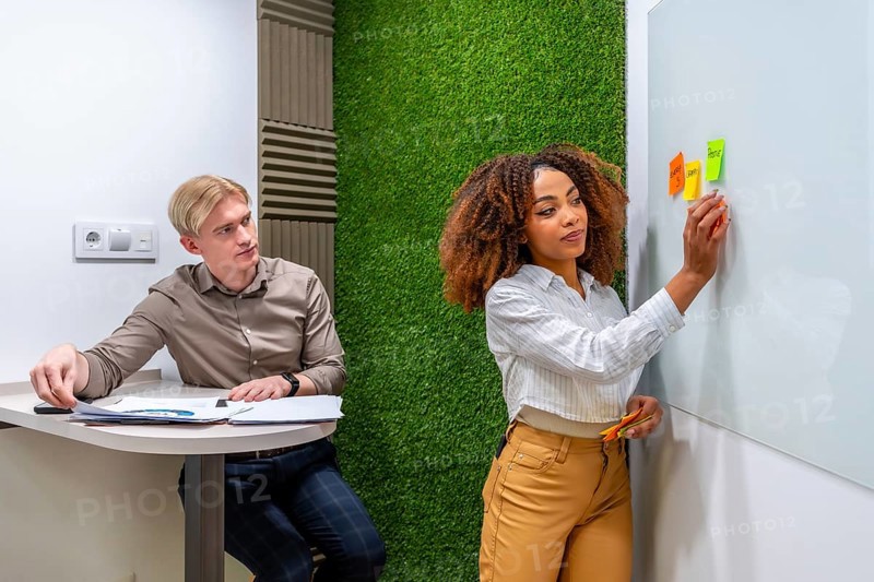 Businesswoman sticking notes on whiteboard while businessman analyzing reports in modern coworking office