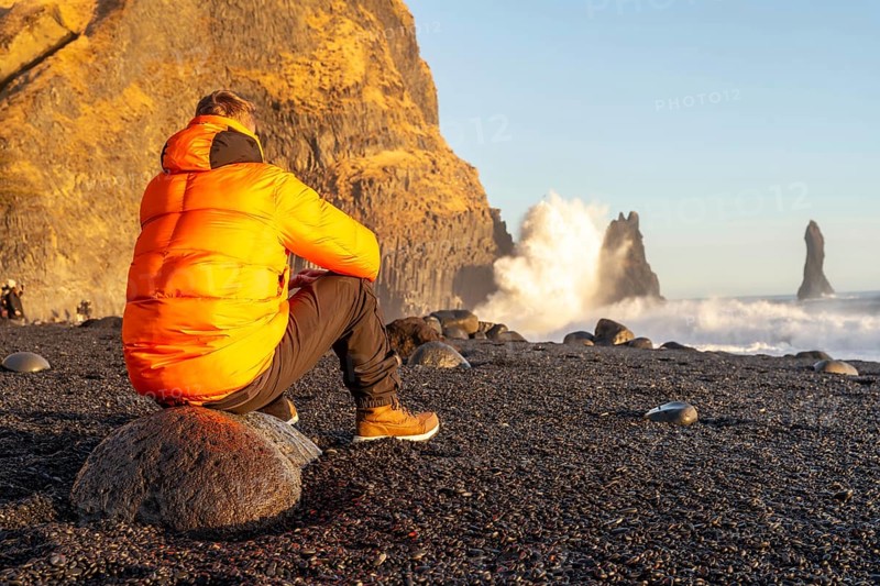 A man sitting on the Reynisfjara Black Sand Beach looking at the beach with a wave