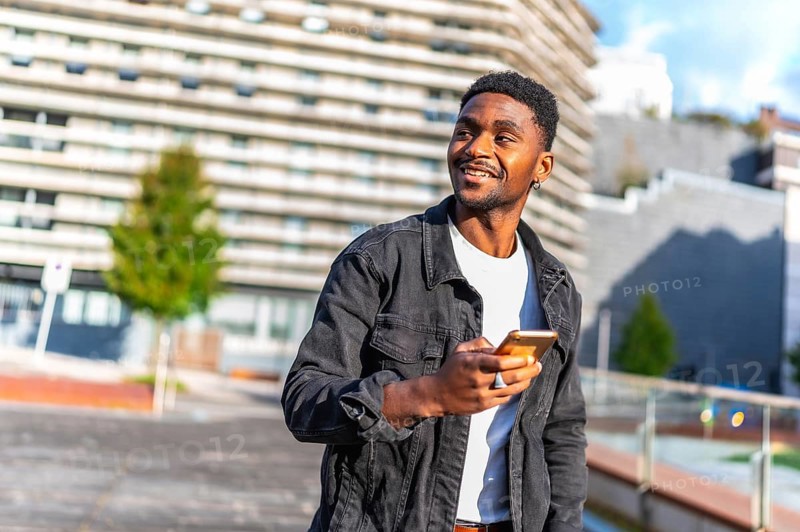 Distracted handsome young african man using mobile app walking along the city