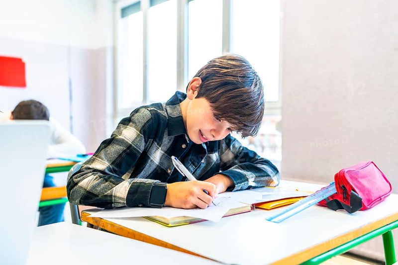 Young boy writing with pen on paper at desk in school classroom, smiling and focused during lesson