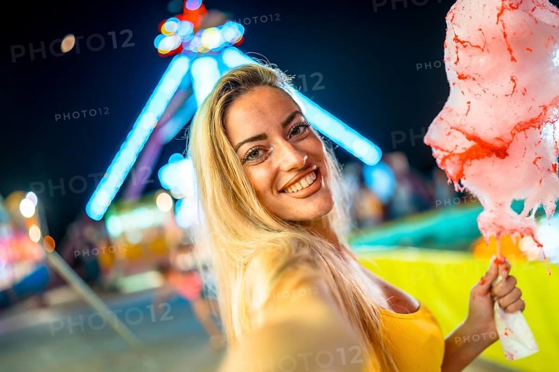 Woman taking selfie while eating pink cotton candy at fair during summer night