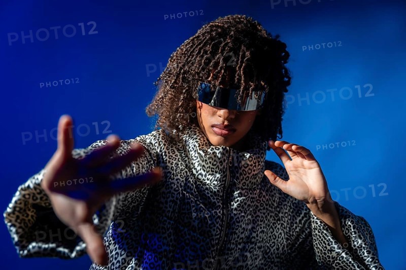 Fashionable woman with curly hair wearing futuristic sunglasses and striking a pose on a vibrant blue backdrop