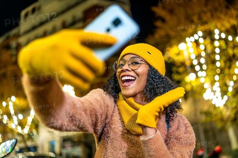 Smiling woman wearing yellow woolen gloves, hat and scarf taking selfie with her smartphone in a city street decorated with christmas lights