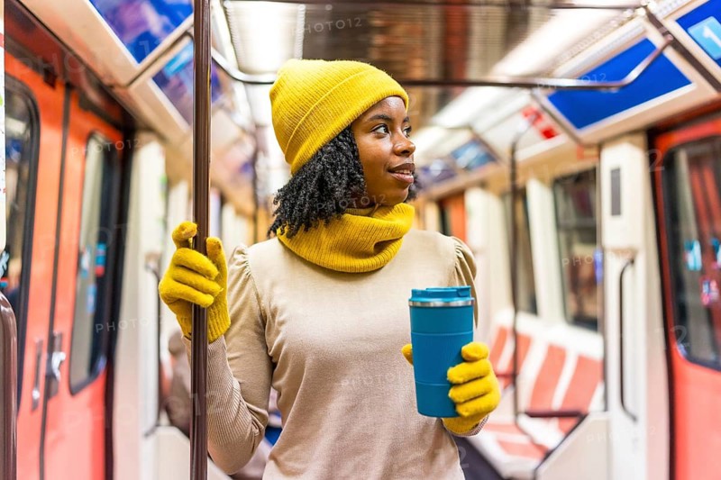 Black woman wearing yellow winter clothes holding a blue reusable coffee cup standing on a subway train