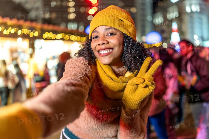 Smiling young woman wearing yellow winter clothes takes a selfie at christmas market showing peace sign with fingers