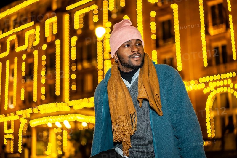 African man sitting outdoors during Christmas night facing Christmas fairy lights decorating a facade