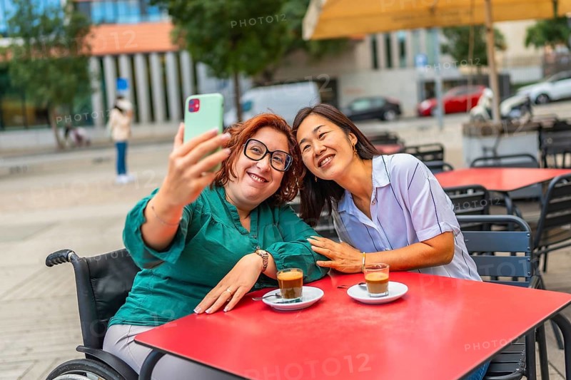 Woman with disability and asian female friend taking selfie in an outdoor cafeteria