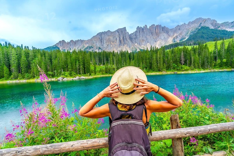 Female hiker with backpack holding her straw hat while admiring the turquoise waters of lake carezza and the stunning latemar peaks in the dolomites