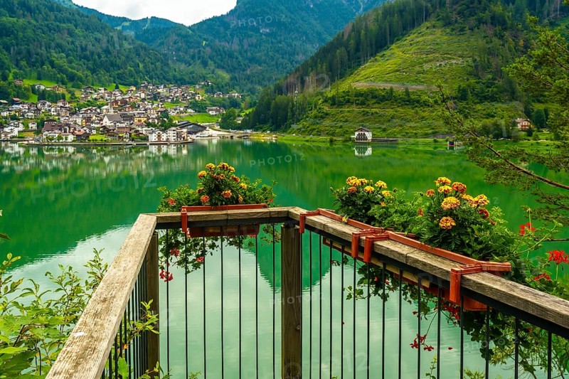 Wooden balcony with flower boxes overlooking the picturesque village of alleghe, reflecting on emerald green lake waters in the dolomites