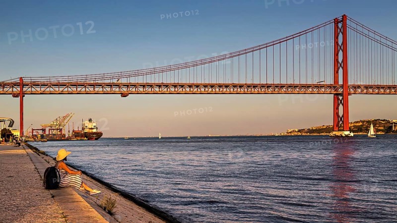 A person sits by a riverbank, gazing at the bridge and boats during a calm sunset, 25 de Abril Bridge, Lisbon