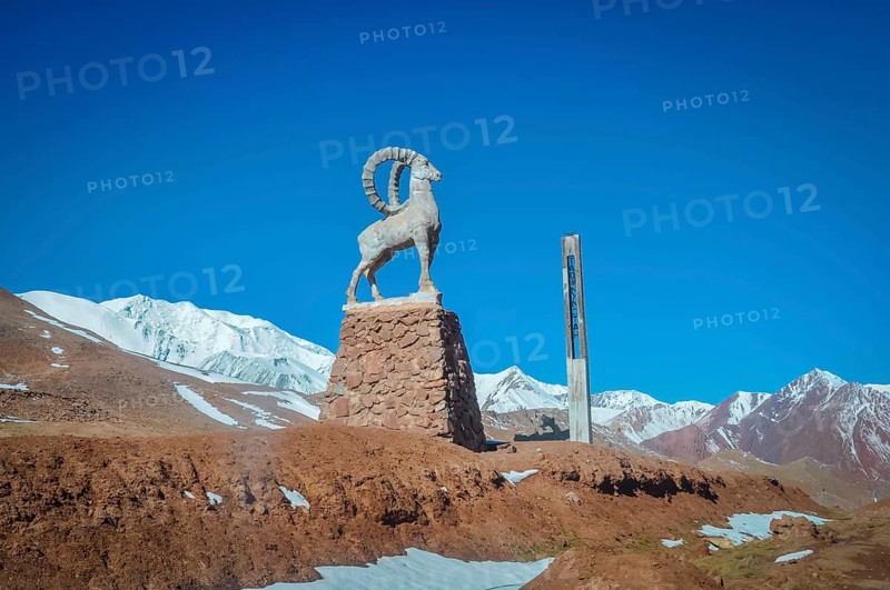 Sculpture of bouquetin on stony base surrounded by mountains with snowy peaks in distance in Karakul in Tajikistan