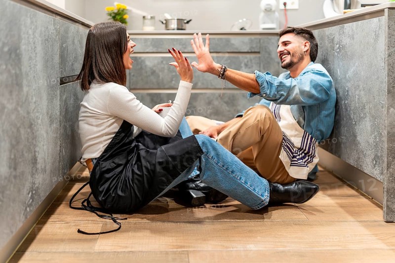 Two young chefs are sitting on the kitchen floor, giving each other a high five and laughing, celebrating their successful work