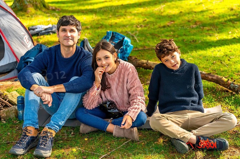 Happy caucasian father, daughter and son sitting next to camping tent enjoying nature in the forest during weekend