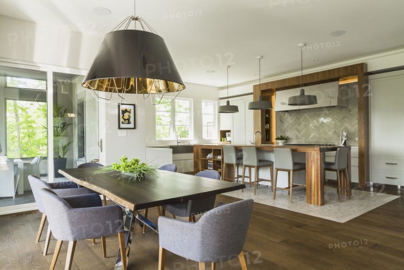 Chrome and walnut wood table with charcoal linen armchairs, large black and gold industrial style pendant lighting fixture and walnut wood island with grey leather and wooden high chairs on travertine mosaic floor surrounded by engineered hickory wood floorboards in dining room kitchen area inside a luxurious contemporary bungalow style home, Quebec, Canada, North America