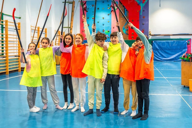 Group of cheerful students wearing colorful bibs raising hockey sticks and celebrating in school gym class